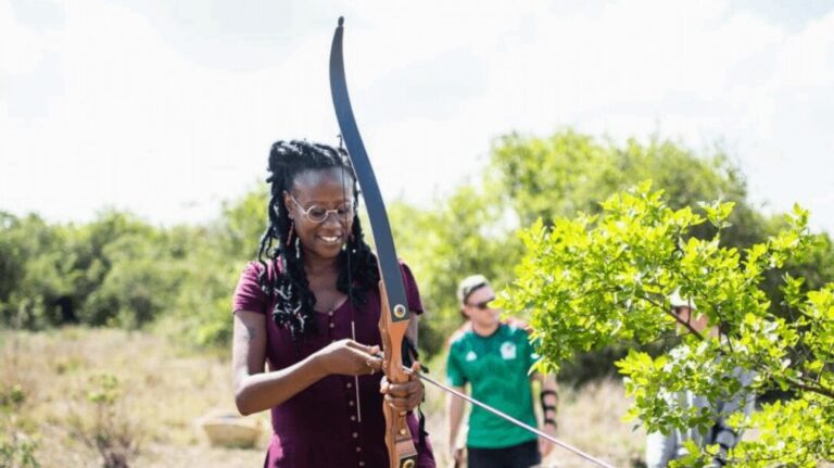 A woman doing archery at Sandai Farm, with a traditional bow in her hand, surrounded by green nature and other participants in the background.